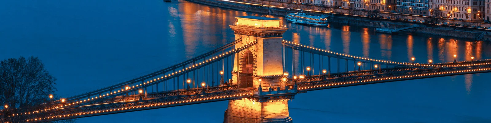 Die Kettenbrücke in Budapest, stimmungsvoll mit warmem Licht beleuchtet, überspannt die Donau. Die Stadtlandschaft spiegelt sich nachts im ruhigen Wasser wider.