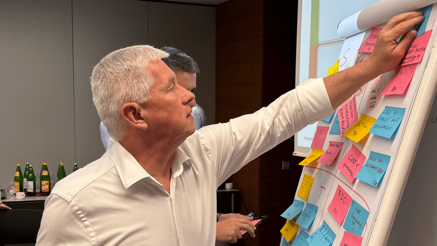 Ein Mann in einem weißen Hemd ordnet in einem modernen Büro bunte Haftnotizen auf einem Flipchart an. Im Hintergrund ist ein Tisch mit Flaschen und Gläsern zu sehen, was auf eine Gruppenarbeit oder ein Brainstorming hindeutet. Die Haftnotizen enthalten handgeschriebenen Text, der einen aktiven Planungs- oder Ideenfindungsprozess signalisiert.