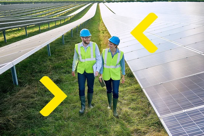 Zwei Ingenieure mit Sicherheitswesten und Helmen gehen durch eine große Solarfarm und bewerten die Effizienz und den Aufbau von Solarmodulen zur Erzeugung erneuerbarer Energie.