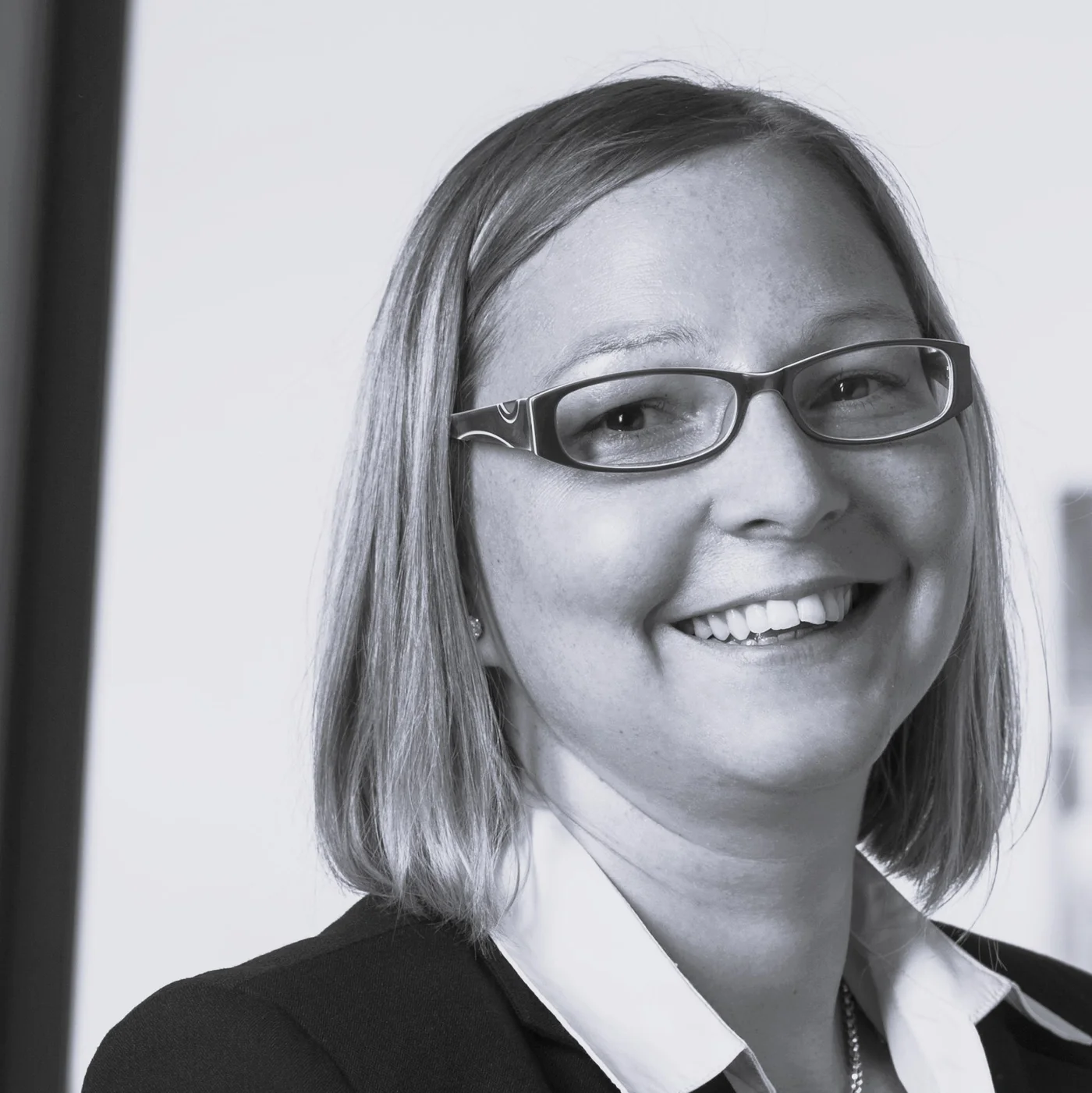 A woman wearing a black blazer and white collared shirt is pictured indoors. The setting appears to be an office or professional environment, with neutral tones and minimal background details. Her hairstyle is a straight bob cut, and the overall mood is formal and business-oriented.