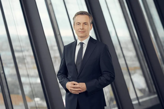 Martin Hauer steht vor den hohen Glasfenstern eines modernen Bürogebäudes. Er trägt einen dunklen Anzug, ein weißes Hemd und eine Krawatte, die Hände locker verschränkt. Im Hintergrund verschwimmt durch die großen Fensterscheiben die Skyline der Stadt. Natürliches Tageslicht schafft eine klare, professionelle Atmosphäre. Die Szene vermittelt Professionalität, Souveränität und Führungsstärke.