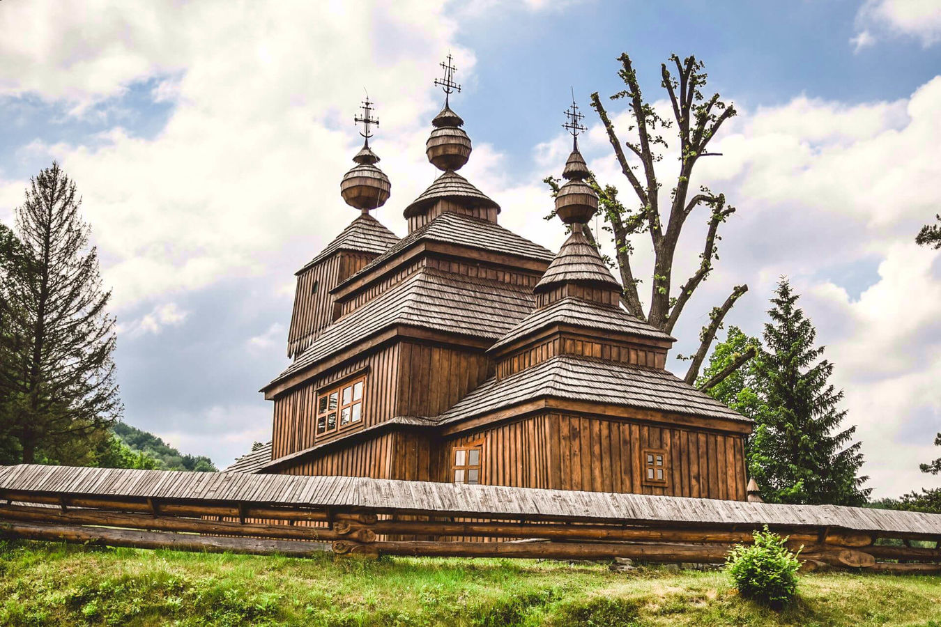 Historische Holzkirche mit mehreren Türmen und Schindeldächern, umgeben von Bäumen und einem Holzzaun.