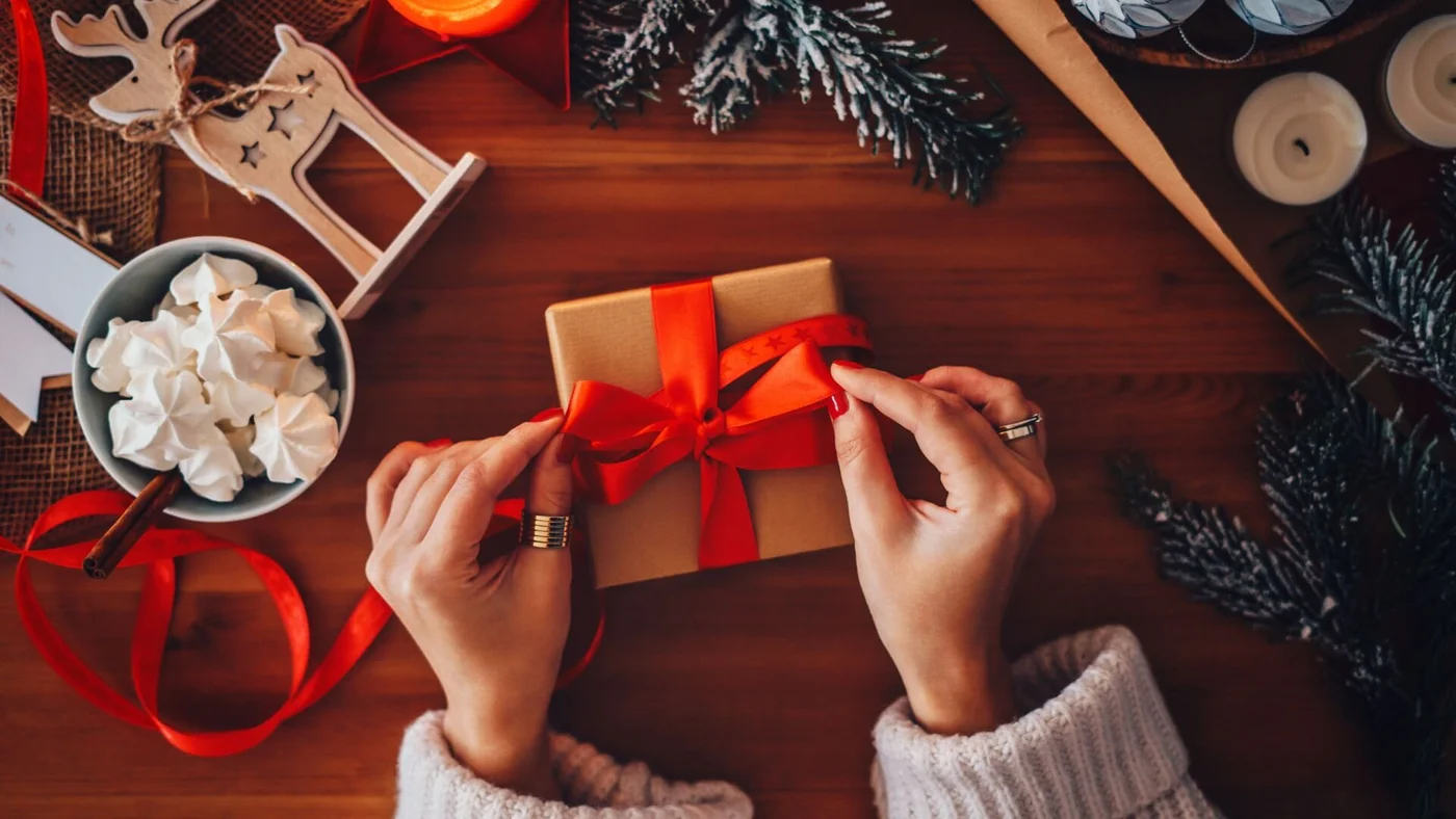 Hände binden ein rotes Band um eine braune Geschenkbox, umgeben von Weihnachtsdekoration, Kerzen und einer Schale mit Baisers.
