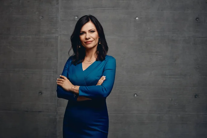 Sladjena Krizaj steht mit verschränkten Armen in einem figurbetonten blauen Kleid vor einer dunkelgrauen Betonwand, die an ein modernes Büro oder Studio erinnert. Ihr Gesicht ist zum Schutz der Anonymität unkenntlich gemacht, doch ihre Haltung strahlt Selbstbewusstsein und Professionalität aus. Dezenter Schmuck und der minimalistische Hintergrund lenken den Fokus auf ihre souveräne Ausstrahlung.
