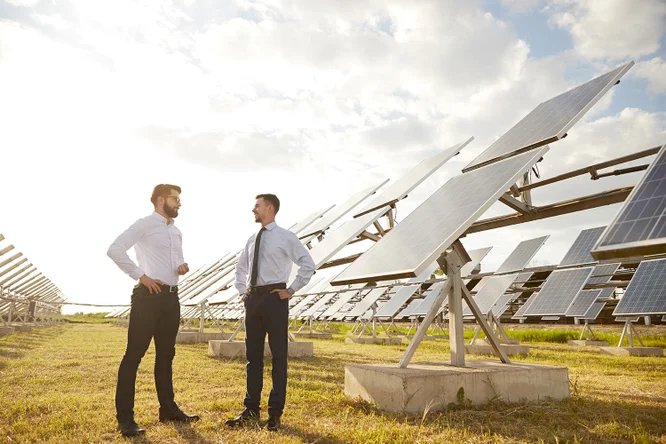 Zwei Geschäftsleute stehen in einem Feld mit Solarpanelen und sprechen über Lösungen für erneuerbare Energien. Die Panele sind zur Sonne hin ausgerichtet, um Sonnenlicht zur Stromerzeugung zu nutzen.