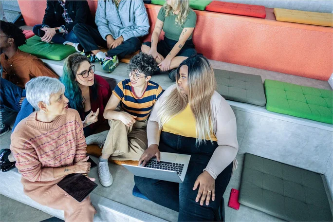 A diverse group of individuals engaged in a collaborative discussion in a modern workspace. The setting features tiered seating with colorful cushions, fostering an open and inclusive environment for sharing ideas.