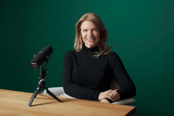 Annette Man sitzt an einem Holztisch mit einem Mikrofon, bereit für eine Solo-Podcast-Aufnahmesitzung vor einem grünen Hintergrund.