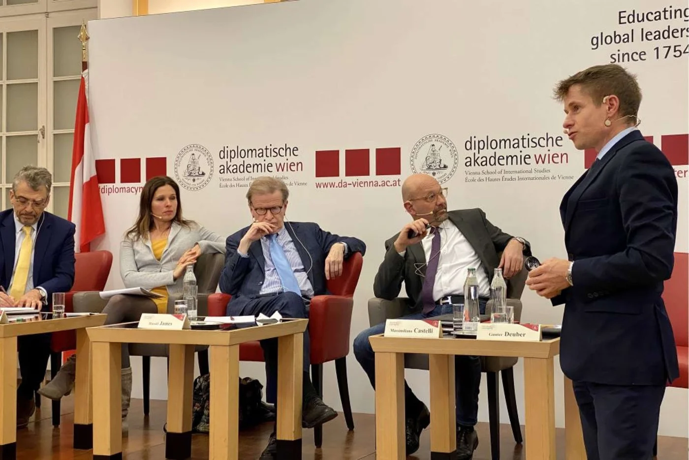 Participants engaged in a panel discussion at the Diplomatic Academy Vienna, seated in red chairs with the Austrian flag in the background. The academy’s branding is prominently displayed.