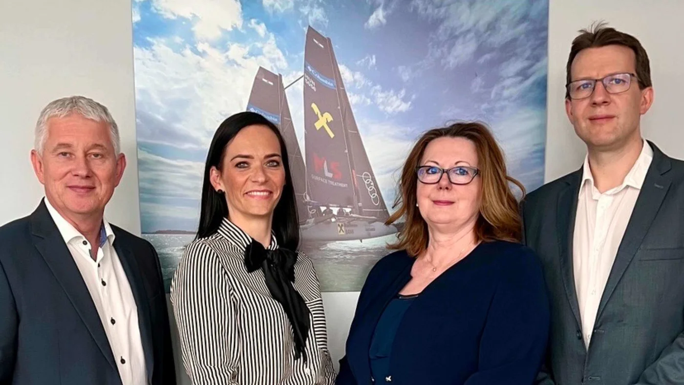 A group of four individuals stands indoors in front of a large sailboat poster. The sailboat features visible text and logos, including 'X' and 'Audi'. The setting appears professional, with formal attire worn by the individuals. The backdrop includes a cloudy sky and ocean visuals, adding depth to the scene.