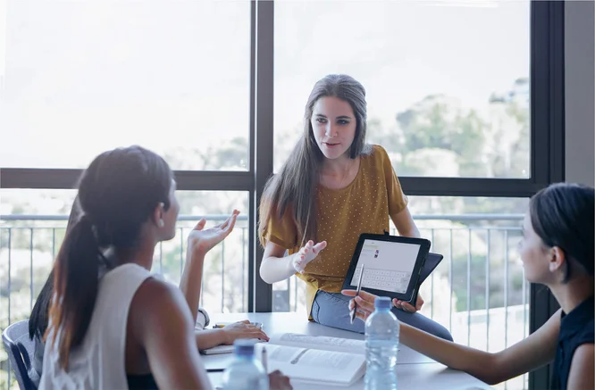 A group of colleagues engaged in a lively discussion in a bright office space, utilizing tablets and documents to facilitate collaboration and idea exchange.