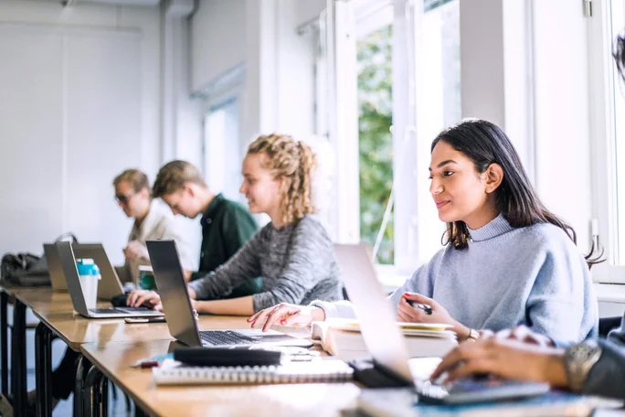 Eine Gruppe von Studenten sitzt an einem langen Tisch und arbeitet mit Laptops und Notizbüchern. Der Klassenraum ist hell, mit großen Fenstern, die natürliches Licht bieten und eine kooperative Lernumgebung fördern.