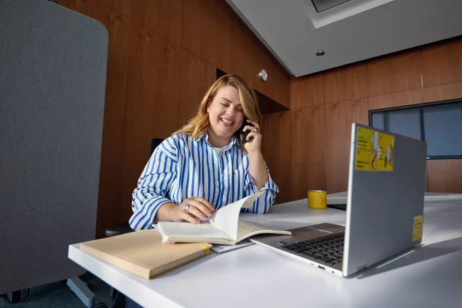 Eine Frau sitzt an einem Schreibtisch in einem modernen Büro und telefoniert, während sie an ihrem Laptop arbeitet. Vor ihr liegen ein offenes Notizbuch und ein geschlossenes Buch, was auf aktives Notizenmachen oder Lernen hindeutet. Der Arbeitsplatz ist mit einer gelben Kaffeetasse und einem Laptop mit gelben Aufklebern ausgestattet, die dem holzgetäfelten Raum einen Farbtupfer verleihen. Die Szene vermittelt insgesamt ein konzentriertes, professionelles Arbeitsumfeld.