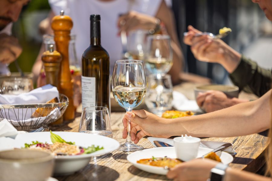 Menschen genießen eine Mahlzeit im Freien mit Tellern voller Essen, Gläsern Weißwein und einer Weinflasche auf einem Holztisch.