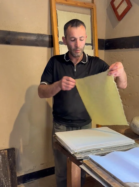 An adult man stands indoors carefully holding a sheet of handmade paper. He is positioned beside a wooden table stacked with neatly arranged paper sheets. The rustic workshop setting features wooden beams, textured walls, and simple tools. Warm lighting emphasizes the texture of the paper and the craftsman’s focused posture.
