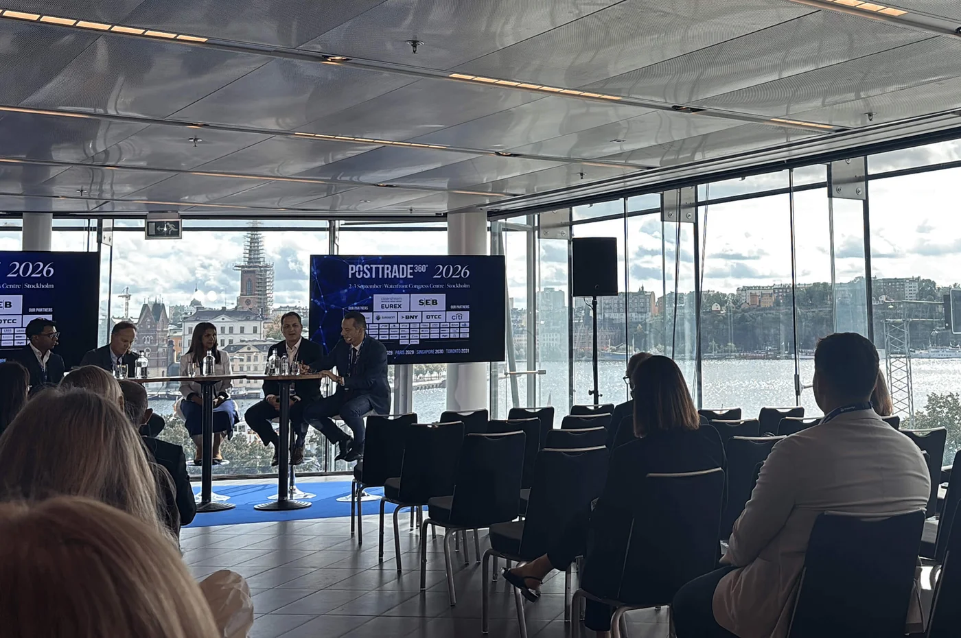 In einem modernen Konferenzraum mit großen Glasfenstern sitzt eine Gruppe von Geschäftsleuten auf einer Bühne zu einer Podiumsdiskussion. Die Teilnehmer sitzen den Rednern zugewandt und verfolgen den Vortrag. Zwei große Bildschirme zeigen den Schriftzug „PSI TRADE™ 2026“ sowie Diagramme und Informationen. Durch die Fenster bietet sich ein Blick auf die Küstenlandschaft und den bewölkten Himmel, wodurch der Raum mit natürlichem Licht erhellt wird.