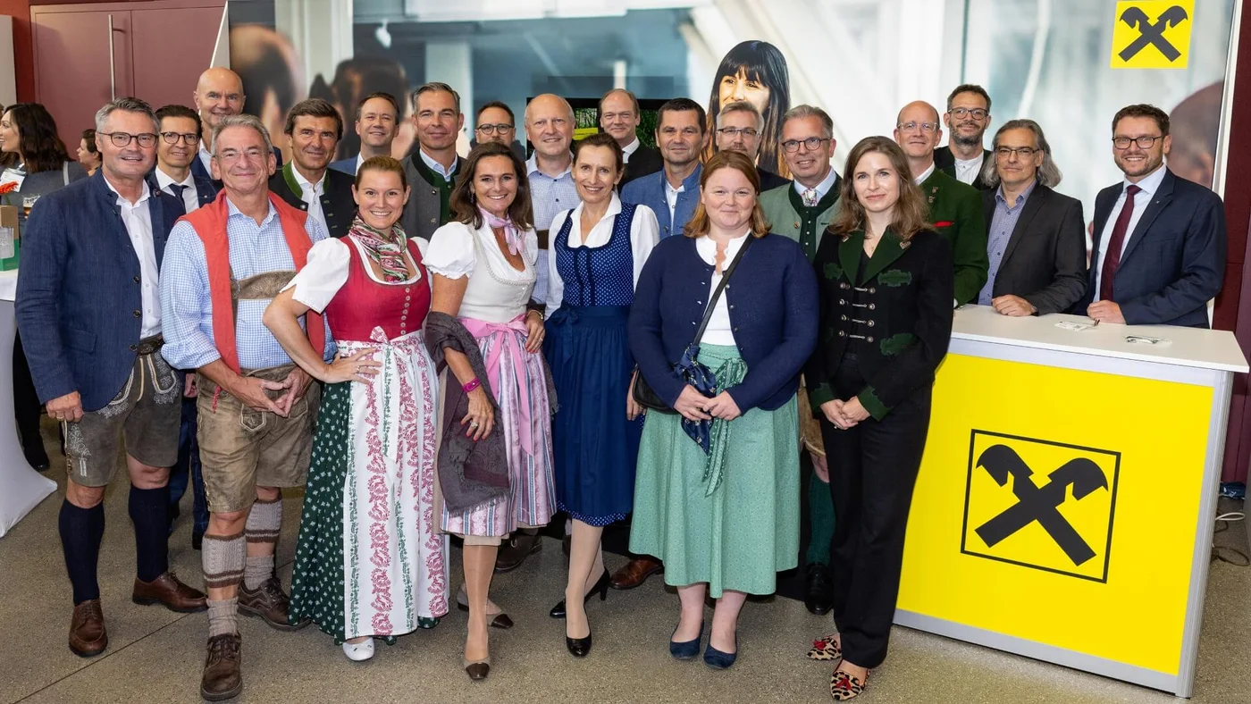 Eine große Gruppe von Menschen versammelt sich in Innenräumen und steht vor einem Stand mit Raiffeisen-Bank-Branding.
