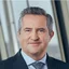 Rainer Schnabl in a suit and tie, looking directly at the camera with a slight smile. He appears in a bright, modern office environment with large windows and soft daylight. The image has a professional, corporate feel with neutral colors and a clean composition. His expression conveys confidence, approachability, and leadership.