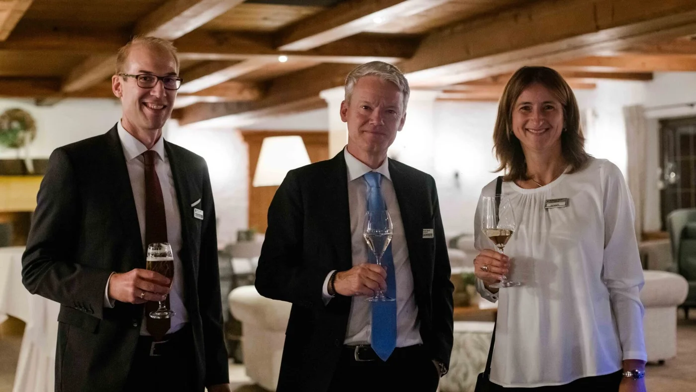 Drei Personen in Geschäftskleidung stehen zusammen und halten Getränke in einem warm beleuchteten Raum mit Holzdecke.