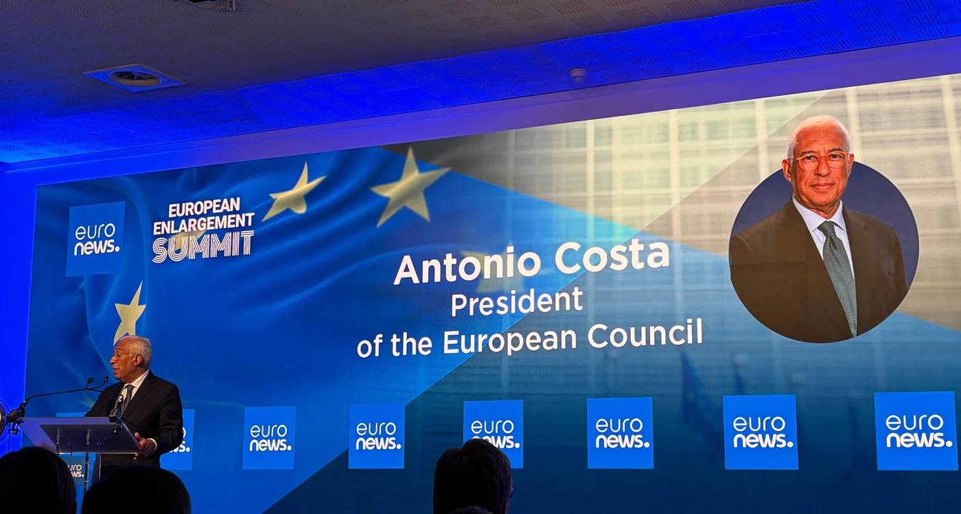 A formal conference stage is set for the European Enlargement Summit. A speaker stands at a podium on the left, addressing the audience in front of a large illuminated backdrop. The backdrop displays the text "EUROPEAN ENLARGEMENT SUMMIT" and "Antonio Costa President of the European Council" alongside a blurred portrait. Multiple blue screens with the "euro news." text are visible across the stage. The overall atmosphere is professional and political, with blue lighting and European flag imagery.