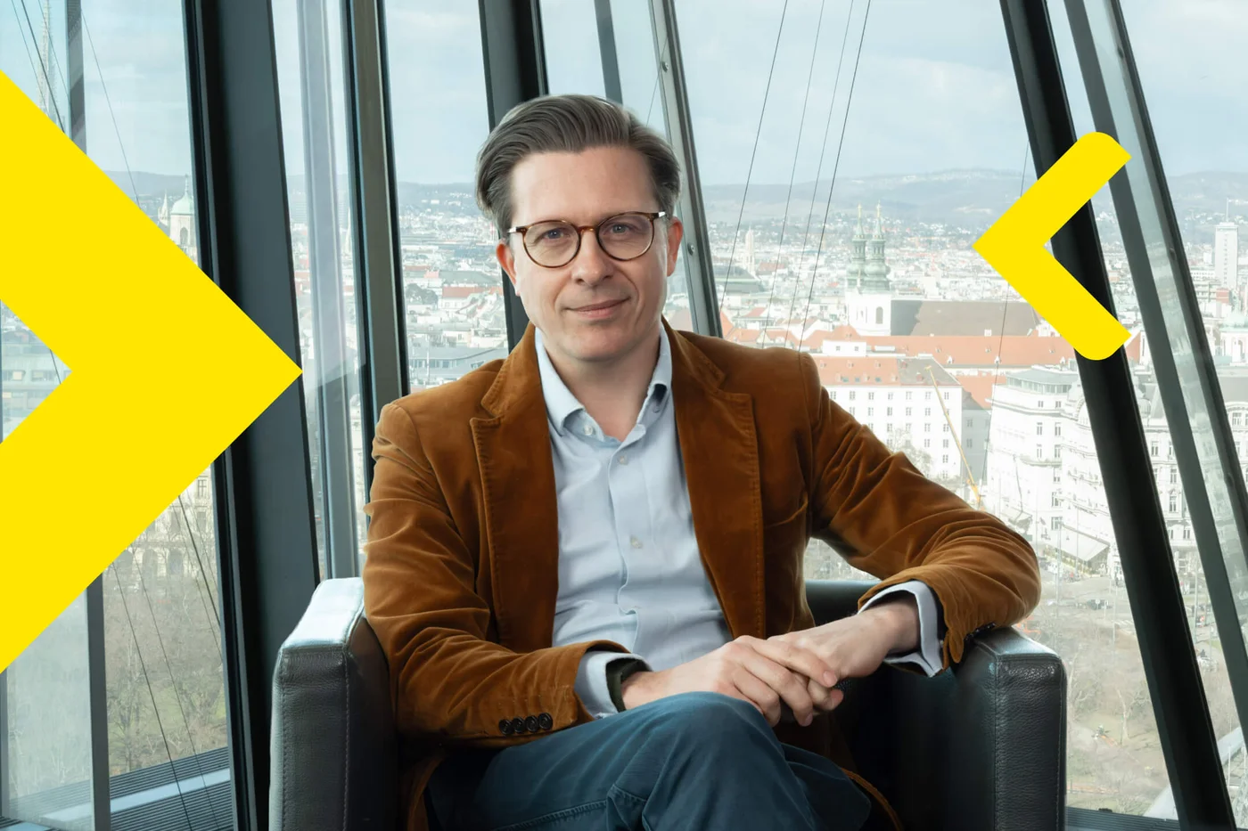 Björn Zaksek wearing a brown blazer and light blue shirt sits in a modern armchair near tall glass windows. The background reveals a cityscape with buildings and distant hills under a cloudy sky. Bright yellow geometric shapes are overlaid on the image, adding a contemporary visual element. The setting appears to be a high-rise or office with panoramic views.
