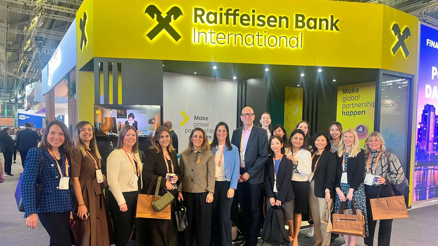 Eine Gruppe von Fachleuten posiert vor dem Stand der Raiffeisen Bank International auf einer Firmenveranstaltung. Der Stand präsentiert sich in leuchtendem Gelb und Schwarz mit dem prominent platzierten Logo der Raiffeisen Bank International. Der sichtbare Text lautet: „Globale Partnerschaften verwirklichen“. Die Veranstaltung findet in einem Ausstellungs- oder Konferenzraum statt.