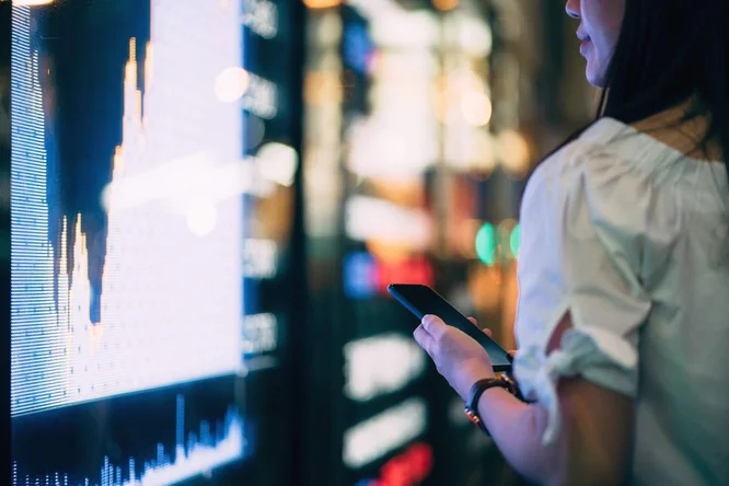 Eine Frau betrachtet Finanzdiagramme auf einem großen digitalen Display und hält dabei ein Smartphone – steht für moderne Aktienmarktanalyse, Handel und Investmenttechnologie.