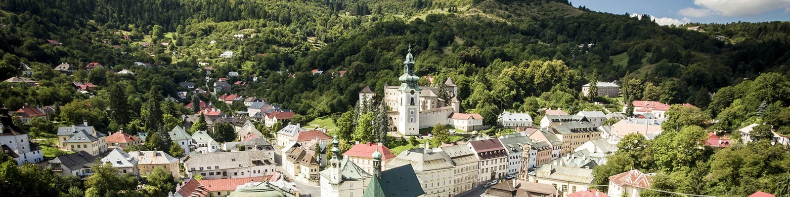 Luftaufnahme einer historischen slowakischen Stadt mit einer markanten Kirche, bunten Dächern und grünen Hügeln im Hintergrund.