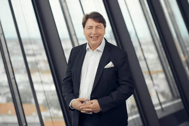 Rudolf Könighofer, in dunklem Anzug und weißem Hemd, steht selbstbewusst vor raumhohen Fenstern. Er lächelt freundlich und verschränkt die Hände – ein professioneller und zugleich zugänglicher Eindruck. Im Hintergrund verschwimmt durch die schrägen Glasscheiben eine urbane Skyline. Die Szenerie erinnert an ein modernes Büroumfeld und ein professionelles Porträt-Shooting.