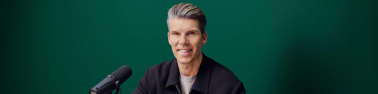 Ein Mann sitzt an einem Schreibtisch vor einem professionellen Mikrofon und lächelt in die Kamera. Die Szene wirkt wie ein Podcast- oder Aufnahmestudio vor einem einfarbig grünen Hintergrund. Er trägt eine dunkle Jacke und hat ordentlich frisiertes graues Haar. Die Bildkomposition ist klar und minimalistisch und konzentriert sich auf den Moderator und die Audioausrüstung.
