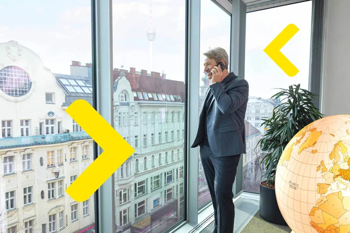 A business professional stands by large windows in a modern office, talking on the phone, with a cityscape view and a large globe in the corner.