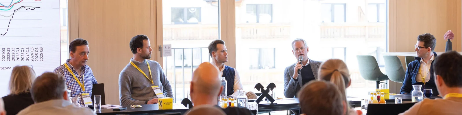 A group of five men are seated at a long table, participating in a panel discussion. The setting is a modern conference room with large windows and natural light. Attendees are visible in the foreground, and a presentation slide with a line graph and numbers is displayed on the left. The atmosphere appears professional and collaborative, with microphones and water glasses on the table.