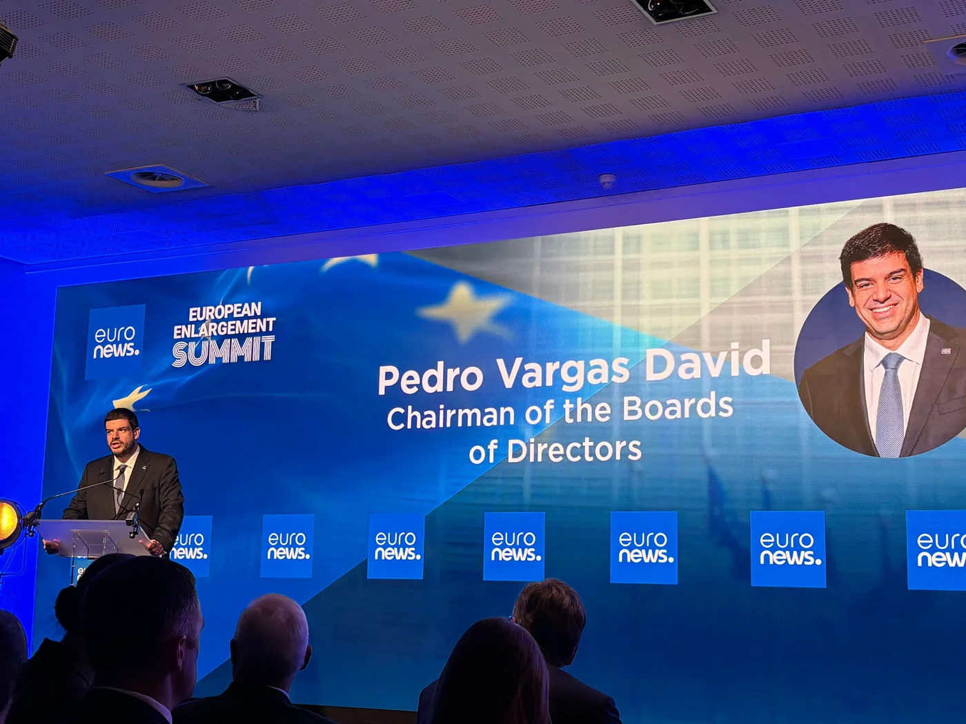 A suited speaker stands at a podium addressing an audience during a formal conference. Behind him, a large illuminated screen displays the text "EUROPEAN ENLARGEMENT SUMMIT" and "Pedro Vargas David Chairman of the Boards of Directors" alongside a portrait of a man in a suit. The stage is lit with blue lighting and features multiple "euro news." panels. Silhouetted attendees are visible in the foreground, emphasizing the professional setting.