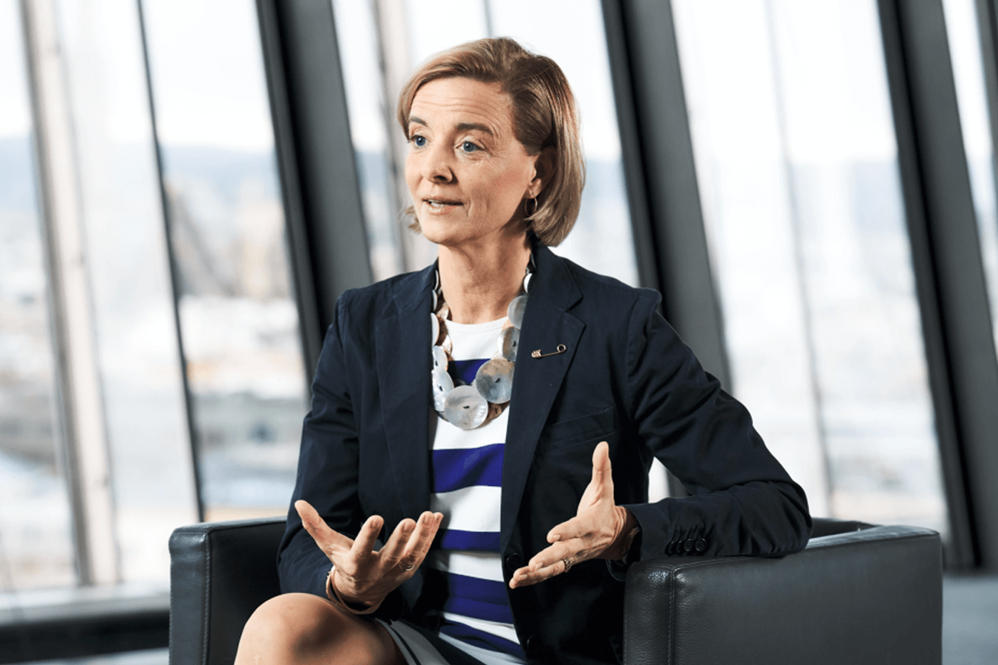 A woman wearing a navy blazer and striped dress is seated in a modern office environment. She gestures with her hands while sitting on a black leather chair. The background features large glass windows with an urban view, creating a professional and contemporary atmosphere.