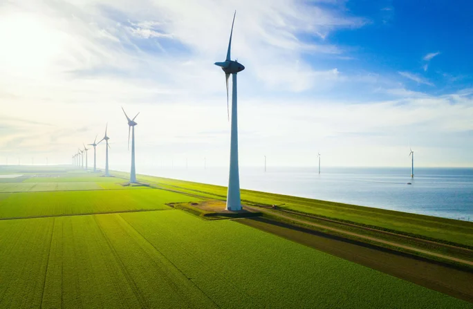 A row of large wind turbines stands tall on expansive green fields adjacent to a calm coastline. The scene is captured under a bright, partly cloudy sky, highlighting the clean energy infrastructure. The landscape features a mix of agricultural land and water, emphasizing sustainable energy production. No visible text or numbers are present in the image.