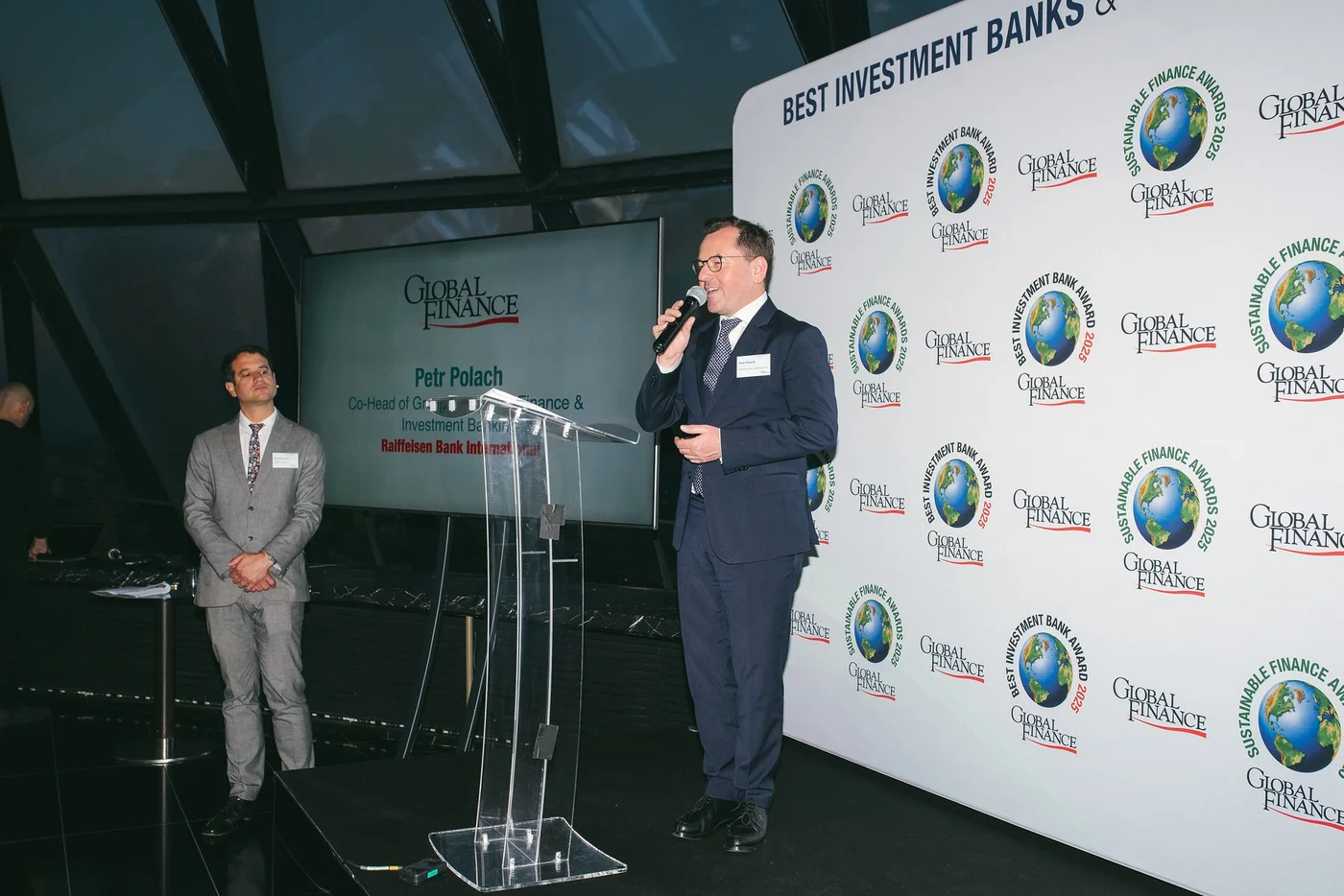 Eine Person im Anzug hält eine Rede am Podium bei den Global Finance Awards vor einer gebrandeten Fotowand und einem Präsentationsbildschirm.