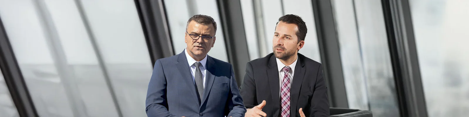 Zwei Personen in formeller Kleidung, die in einem modernen Büro mit großen Fenstern ein Gespräch führen.
