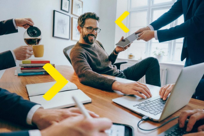 In einem hellen, modernen Büro sitzen Erwachsene an einem Holztisch und arbeiten gemeinsam. Eine Person sitzt mit einem Laptop da, eine andere schenkt Kaffee ein und eine dritte reicht ein Tablet. Die Atmosphäre ist dynamisch und produktiv; sichtbare Notizblöcke, Stifte und digitale Geräte tragen zum professionellen Ambiente bei. Gelbe Pfeile sind zur Verdeutlichung in das Bild eingeblendet.
