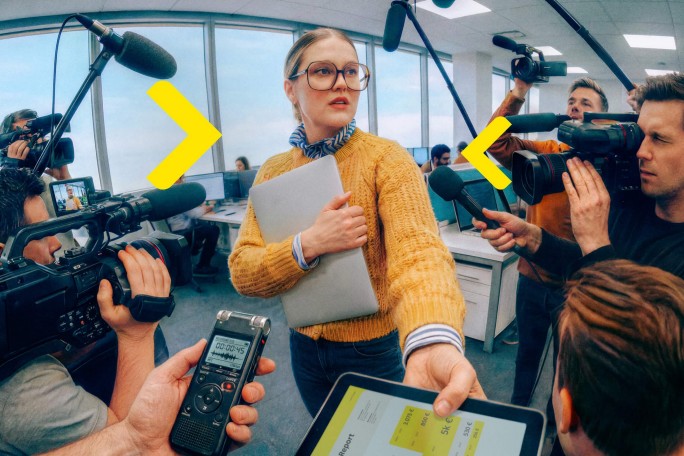 A woman in a yellow sweater holds a laptop while being approached by several reporters with microphones, cameras, and a voice recorder in a modern office setting. The scene is dynamic, with journalists capturing audio and video, and a tablet displaying text in the foreground. The atmosphere is intense and focused, with bright natural light from large windows. No visible brands or explicit numbers are present.