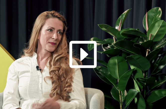 A person with long hair sits in a light-colored armchair during an interview or talk. They wear a white shirt and have a small microphone clipped near the collar. A large green houseplant stands to the right, adding natural contrast against a dark curtain backdrop. A white play button icon is overlaid in the center, indicating this is a video thumbnail. (opens in a new tab)