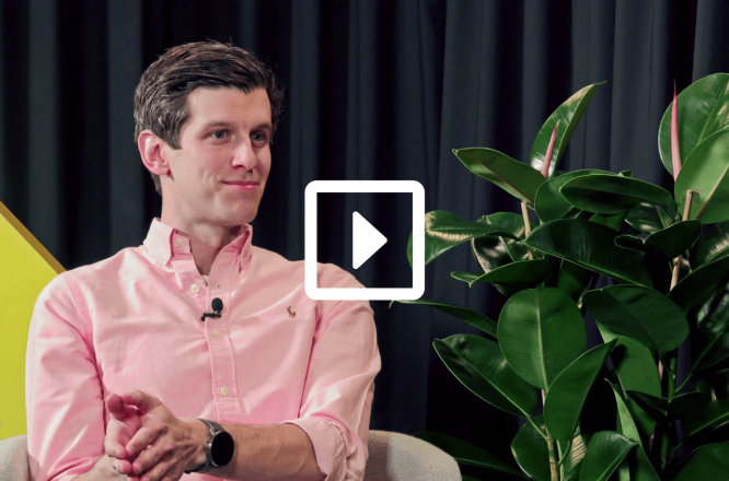 A man wearing a light pink button-down shirt sits in a modern studio setting, partially obscured by a video play icon overlay. He has a small clip-on microphone attached to his shirt and a smartwatch on his wrist, suggesting he is being interviewed or recorded. Behind him is a dark curtain backdrop and a large green houseplant, adding a casual yet professional atmosphere. The composition suggests a recorded talk, podcast, or video interview session. (opens in a new tab)
