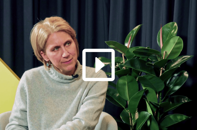 A person sits in a light-colored sweater during an interview or talk show, with their face obscured for privacy. They are seated on a light chair in front of dark curtains, creating a studio-like setting. A large green houseplant with glossy leaves stands prominently to the right. A white play button icon is overlaid in the center, indicating that this is a video thumbnail. (opens in a new tab)