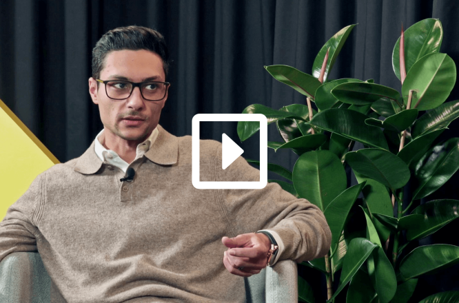 A man sits in a modern studio setting, partially obscured by a play button icon indicating video content. He wears a light brown sweater over a collared shirt and rests comfortably in an armchair. A large green houseplant stands to his side against a dark curtain backdrop, adding a natural element to the scene. The composition suggests a professional interview or talk show environment. (opens in a new tab)