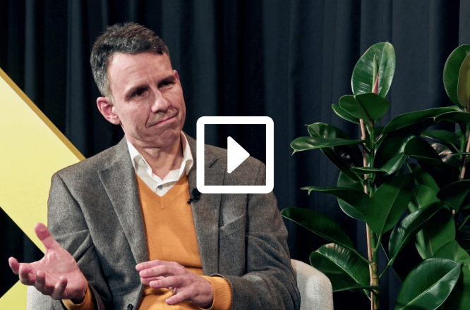 A person in a blazer and sweater sits in a chair during an interview or talk show. The speaker’s face is blurred, and they gesture with one hand while speaking. A large yellow geometric shape stands in the background against dark curtains. A green potted plant is visible to the side, adding a touch of nature to the studio setting. A play button overlay suggests this is a video thumbnail or recording. (opens in a new tab)