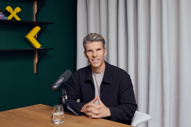 Ein Mann sitzt an einem Holztisch in einem modernen Tonstudio, mit einem vor ihm positionierten Mikrofon in die Kamera gewandt. Auf dem Tisch steht ein Glas Wasser, und geometrische gelbe Wanddekorationen heben sich vom dunkelgrünen Hintergrund ab. Hellfarbene Vorhänge verdecken die Wand hinter ihm und schaffen eine saubere und professionelle Atmosphäre. Das Gesamtbild lässt an eine Podcast- oder Interviewaufnahme in einem modernen, gut beleuchteten Raum denken.