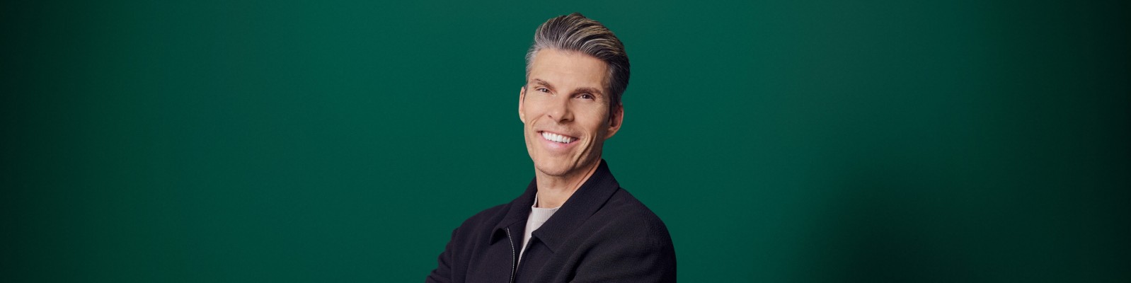 A man with neatly styled gray hair smiles confidently at the camera. He is wearing a dark jacket over a light shirt, giving a polished and professional appearance. The background is a solid green color, creating a clean and modern studio look. The composition focuses on his upper body and friendly expression.