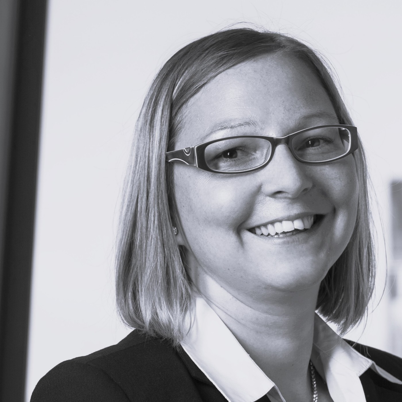 Margit Hybek trägt ein grau kariertes Oberteil und sitzt in einem modernen Büroflur. Im Hintergrund sind Glastüren und ein zeitgemäßes Design mit grünen Akzenten zu sehen. Die Umgebung deutet auf ein professionelles Arbeitsumfeld hin und betont die Ästhetik des Arbeitsplatzes.