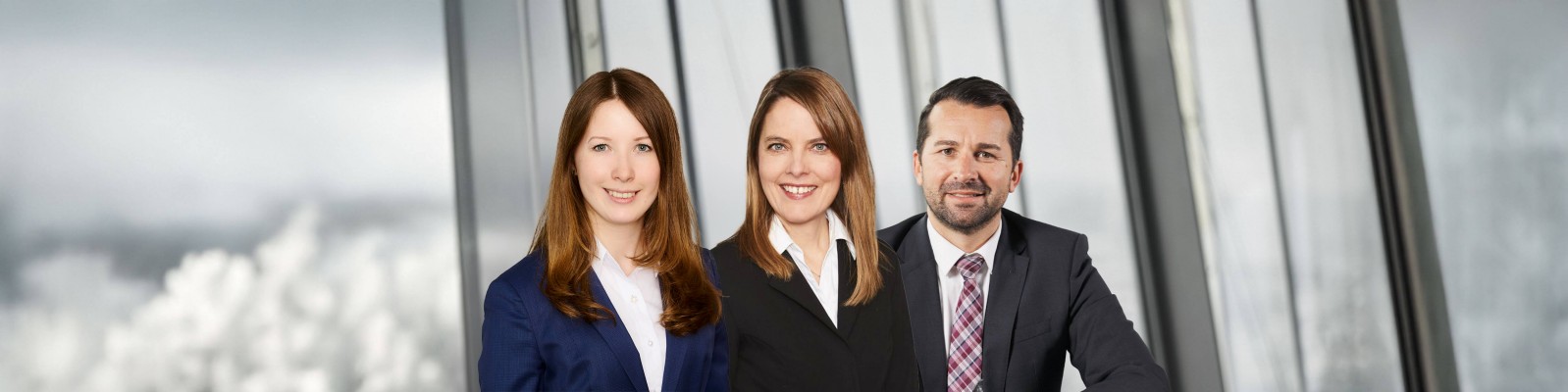 Drei Berufstätige in Business-Anzügen stehen in einem modernen Büro mit großen Fenstern und Blick auf die Stadt.