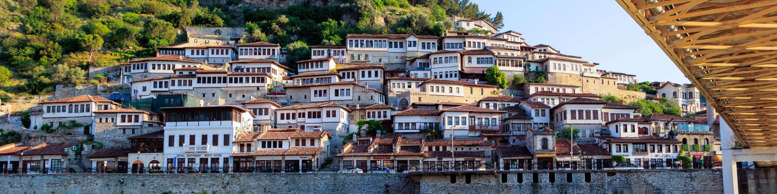 Malerischer Blick auf eine Hügellandschaft mit traditionellen Häusern im osmanischen Stil, weißen Fassaden und roten Dächern. Die Szenerie ist von üppigem Grün umgeben, im Vordergrund befindet sich eine Brücke, die dem Bild Tiefe verleiht. Das Bild fängt den Charme und die historische Atmosphäre des Ortes bei klarem Tageslicht ein.
