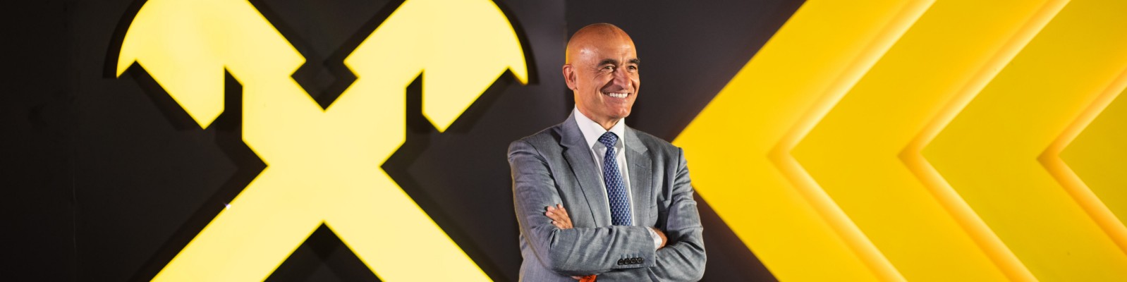 A man in formal attire stands confidently against a striking backdrop featuring bold yellow graphics. The design includes a large crossed hammer symbol and arrow-like shapes, creating a dynamic and professional atmosphere. The setting suggests corporate branding or a business-related event.