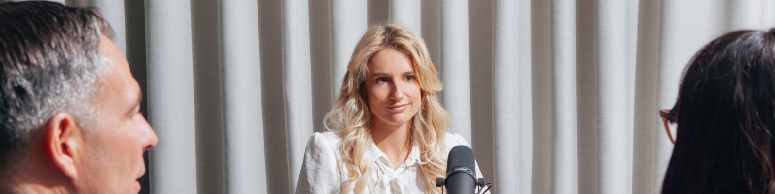 Anna Gasser spricht bei einer formellen Veranstaltung und steht vor einem Mikrofon. Im Hintergrund sind neutrale Vorhänge zu sehen, was auf einen Innenraum hindeutet. Im Vordergrund sind zwei Personen teilweise sichtbar, die aufmerksam zuhören.